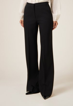 Zwarte wijde broek van gladde stof, met zijzakken en een op maat gemaakte tailleband, gecombineerd met puntige zwarte hakken.