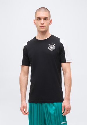 Giovane uomo con una maglia a maniche corte bianca e nera con emblema e pantaloncini Adidas turchese a righe, in piedi davanti a uno sfondo semplice.