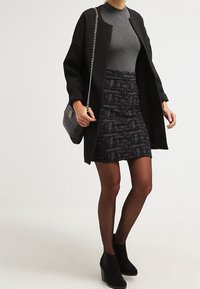 Schwarzer strukturierter Mantel über einem grauen Oberteil, kombiniert mit einem dunkel gemusterten Minirock, schwarzen Strumpfhosen und klobigen, mit Absatz versehenen Stiefeletten. Enthält eine Schultertasche.