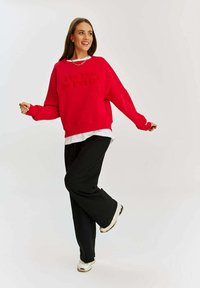 Červená oversized mikina s textúrovaným písmom, vrstvená cez biele tričko, kombinovaná s voľnými čiernymi nohavicami a bielymi teniskami.