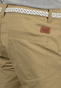 Pantaloni scurți cargo bej, realizati dintr-un material texturat, cu o buzunar la spate și o curea țesută cu un model albastru și alb. Pune eticheta din piele deasupra buzunarului.
