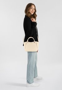 Borsa a mano crema con doppi manici, texture in pelle liscia e tracolla removibile. Il modello indossa un blazer nero e jeans azzurri.