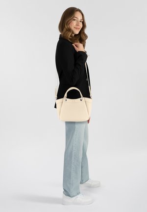 Borsa a mano crema con doppi manici, texture in pelle liscia e tracolla removibile. Il modello indossa un blazer nero e jeans azzurri.