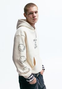 Roomkleurige capuchontrui met de tekst "SMELLS LIKE TEEN SPIRIT". Voorzien van smileygezichtgrafieken op de mouwen, de voorkantzak en geribbelde manchetten.