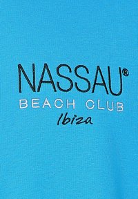 Himmelblaues Tuch mit gesticktem Text: "NASSAU" in Schwarz, "BEACH CLUB" in Weiß und "Ibiza" in geschwungener Schrift in Schwarz am unteren Rand.