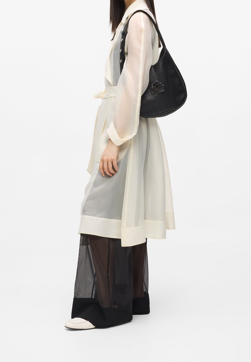 Femme portant un manteau transparent blanc cassé avec un pantalon large noir transparent et portant un sac à bandoulière en cuir noir avec un logo argenté.