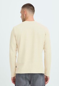 Jasno-beżowa koszula z długim rękawem o teksturowanym wzorze dzianiny i okrągłym dekolcie; posiada małą metkę na dole i luźny krój.