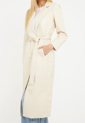 Kvinde iført en lang, cremefarvet frakke med bælte over blå jeans og en hvid top, med den ene hånd i lommen.