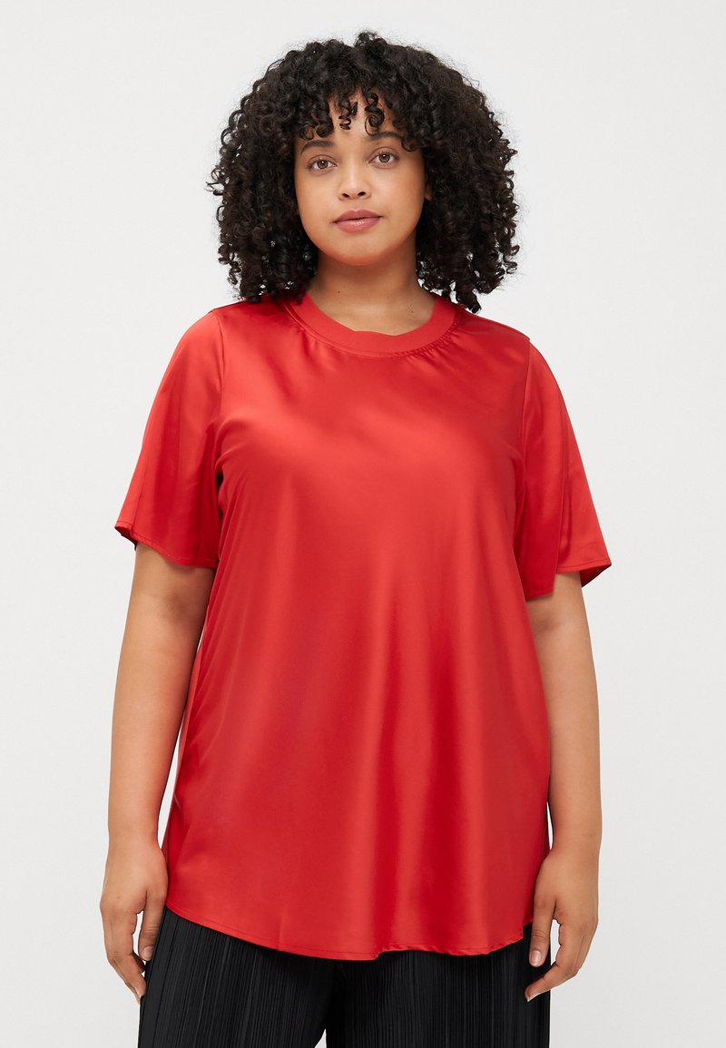 A person with curly hair, wearing a red short-sleeve top, stands against a light background, looking directly at the viewer.