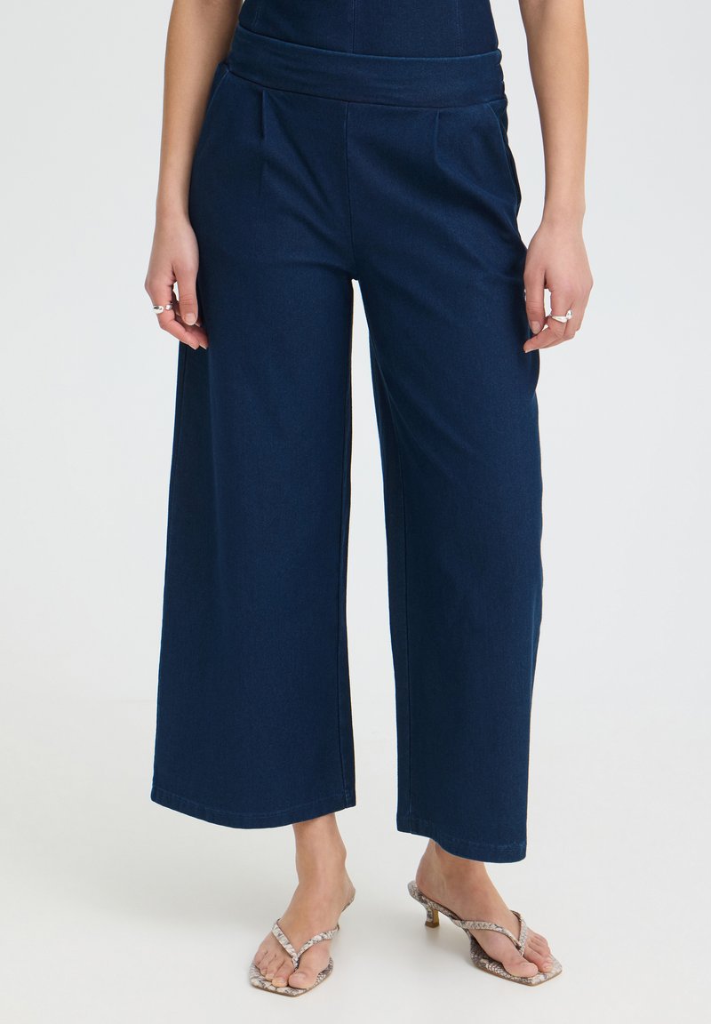 Femme portant un pantalon large bleu marine et des sandales à talons à motif peau de serpent sur un fond uni.