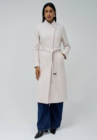 Cappotto lungo rosa chiaro con vita aderente e cintura coordinata. Presenta un colletto alto e una chiusura a singolo bottone. Texture liscia, design su misura.