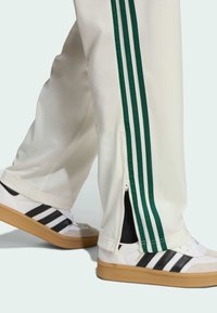 Vita trackbyxor med mörkgröna ränder och sidoglator, i kombination med vita sneakers med svarta ränder och en gummisula.