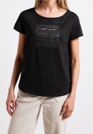 Czarny T-shirt z krótkim rękawem z białym, nakrapianym kwadratem i napisem "zainspiruj się", noszony przez stojącą osobę w jasnobeżowych spodniach.