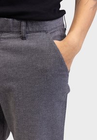 Graue gemusterte Hose mit strukturierter Oberfläche, ausgestattet mit einem Standardbund und Seitentasche, teilweise sichtbare Hand in der Tasche.