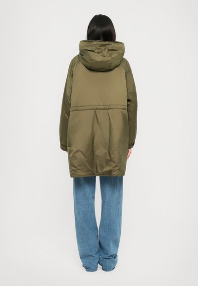Person seen from the back wearing an oversized olive green hooded parka and loose light blue jeans standing against a plain white background.