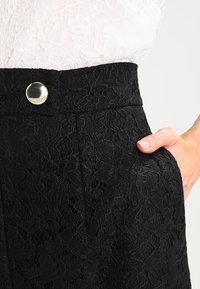 Černé šortky z krajky s texturovaným květinovým vzorem, s viditelnou boční kapsou a kovovým knoflíkovým detailem na pásku.