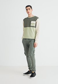 Sudadera en bloque de color verde oliva y crema con un bolsillo delantero, combinada con pantalones cargo. Zapatillas negras completan el conjunto.
