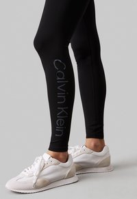 Zwarte leggings hebben een glanzend 'Calvin Klein' logo langs de zijkant. Het model draagt witte sneakers met een gestructureerde zool en zichtbare veters.