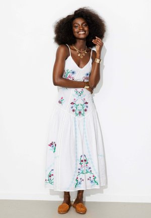 Femme souriante avec un grand afro, portant une robe blanche sans manches brodée de fleurs et des chaussures marron tissées sans lacets, debout contre un mur blanc.