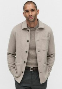 Beige ullblandad jacka med krage, svarta knappar och två framfickor. Bärs över en strukturerad grå tröja, kombinerad med mörka byxor.