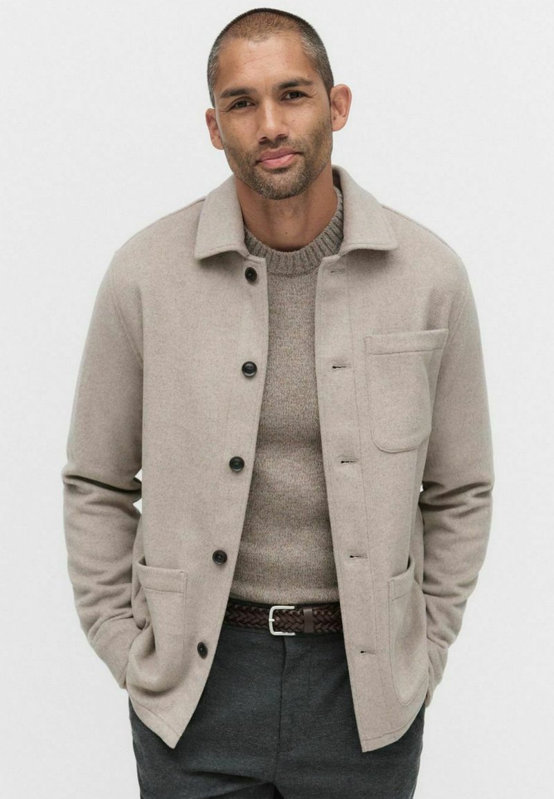 Beige ullblandad jacka med krage, svarta knappar och två framfickor. Bärs över en strukturerad grå tröja, kombinerad med mörka byxor.