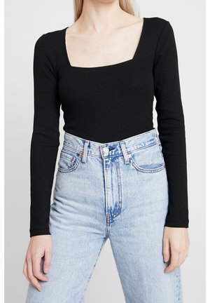 Vrouw draagt een zwarte top met lange mouwen en een vierkante hals, ingestopt in een lichtblauwe spijkerbroek met hoge taille, tegen een effen achtergrond.
