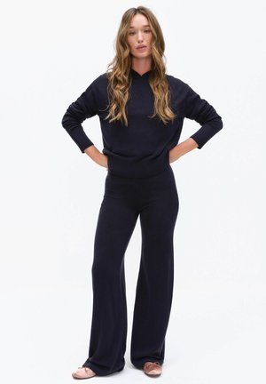 Donna con capelli lunghi e mossi in piedi con le mani sui fianchi, indossa un maglione blu navy, pantaloni a gamba larga e ballerine rosa su sfondo bianco.