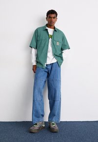 Green short-sleeve shirt over a white long-sleeve shirt, paired with wide blue jeans and gray sneakers with green accents.