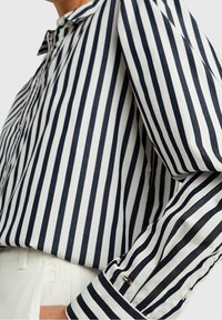 Camisa de botones con rayas verticales en navy y blanco, que cuenta con un cuello, mangas largas y un pequeño logo en el puño.
