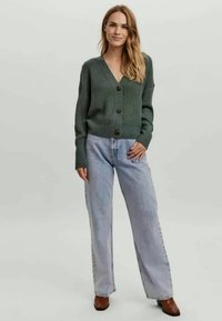 Zelený žebrovaný cardigan s výstřihem do V, s třemi velkými knoflíky, kombinovaný se světlomodrými širokými džíny a hnědými botami na podpatku.