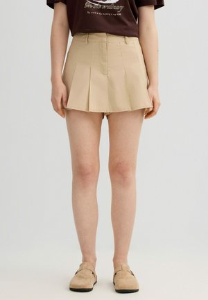 Mujer que lleva una falda-pantalón plisada beige combinada con camisa marrón y zapatos beige sin cordones, de pie frente a un fondo claro y liso.