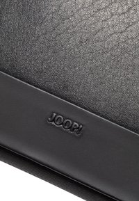 Materiale di pelle nera texture con logo "JOOP!" in rilievo su una striscia liscia, caratterizzato da un sottile motivo a grana fine.