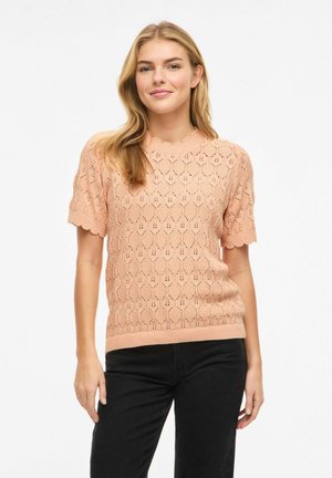 Mlada ženska, oblečena v breskvasto majico s kratkimi rokavi z vzorcem v teksturi in črnimi hlačami, stoji pred belim ozadjem.