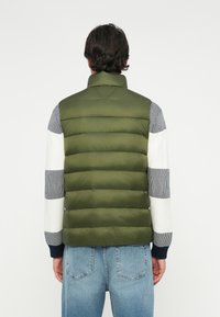 Vestă căptușită de culoare verde măslinie, cu guler înalt, având secțiuni cusute orizontal. Este purtată peste o bluză cu dungi multiple și mâneci lungi, împreună cu blugi de culoare bleu deschis.