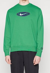 Grön sweatshirt med långa ärmar, ribbade muddar och fåll. Har en marinblå oval logotyp med "NIKE" i grönt på bröstet. Mjuk textur.