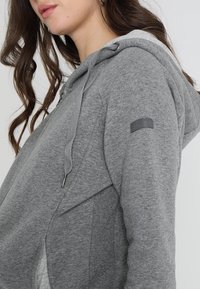 Donna che indossa una felpa grigia con zip e cappuccio regolabile, posata in una vista laterale ravvicinata che mostra spalla e parte del viso.