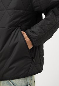 Sort quiltet jakke med en struktureret overflade, der har en sidelomme og elastiske manchetter. Designet fremhæver et geometrisk diamantmønster.