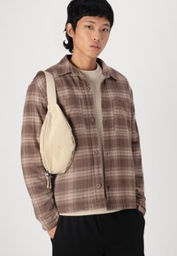 Borsa a tracolla beige con texture liscia, portata sulla spalla; il modello indossa una camicia a quadri marrone sopra un maglione chiaro.