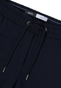 Donkerblauwe broek met een elastische tailleband, een trekkoord in donkerblauw met metalen tips, en zijzakken. Zachte stof met een gladde textuur.