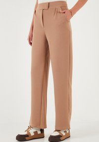 Beige leveled-leghousut, jotka on valmistettu tekstuurikankaasta, korkea vyötärö, etuompelukset ja sivutaskut, yhdistettynä paksuihin lenkkareihin.