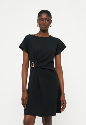 Γυναίκα που φοράει μια μαύρη κοντομάνικη φορέμα με λεπτομέρεια χρυσής αγκράφας στη μέση, στέκεται μπροστά σε ένα απλό λευκό φόντο.