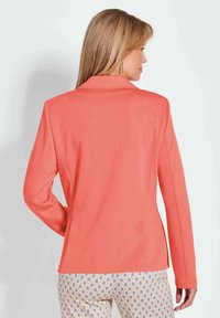 Koraalblazer met een gladde textuur, gestructureerde schouders en een getailleerde pasvorm. Opvallende lange mouwen en een standaard reverskraag, gecombineerd met gestreepte broek.