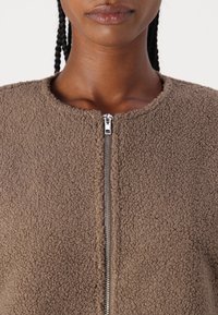 Detailní z�áběr na osobu oblečenou v hnědé strukturované fleecové bundě se středovým stříbrným zipem a kulatým výstřihem.
