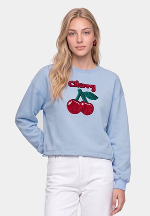 Kvinde med langt blondt hår iført en lyseblå sweatshirt med røde kirsebær og ordet "Cherry" i røde palietter, kombineret med hvide bukser.