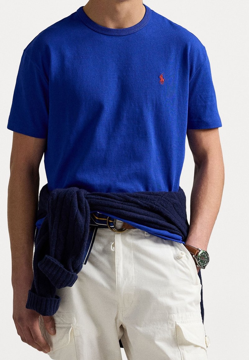 Camiseta de algodón azul real con cuello redondo acanalado, con bordado de logo rojo en el pecho. Cintura visible con pantalones blancos.