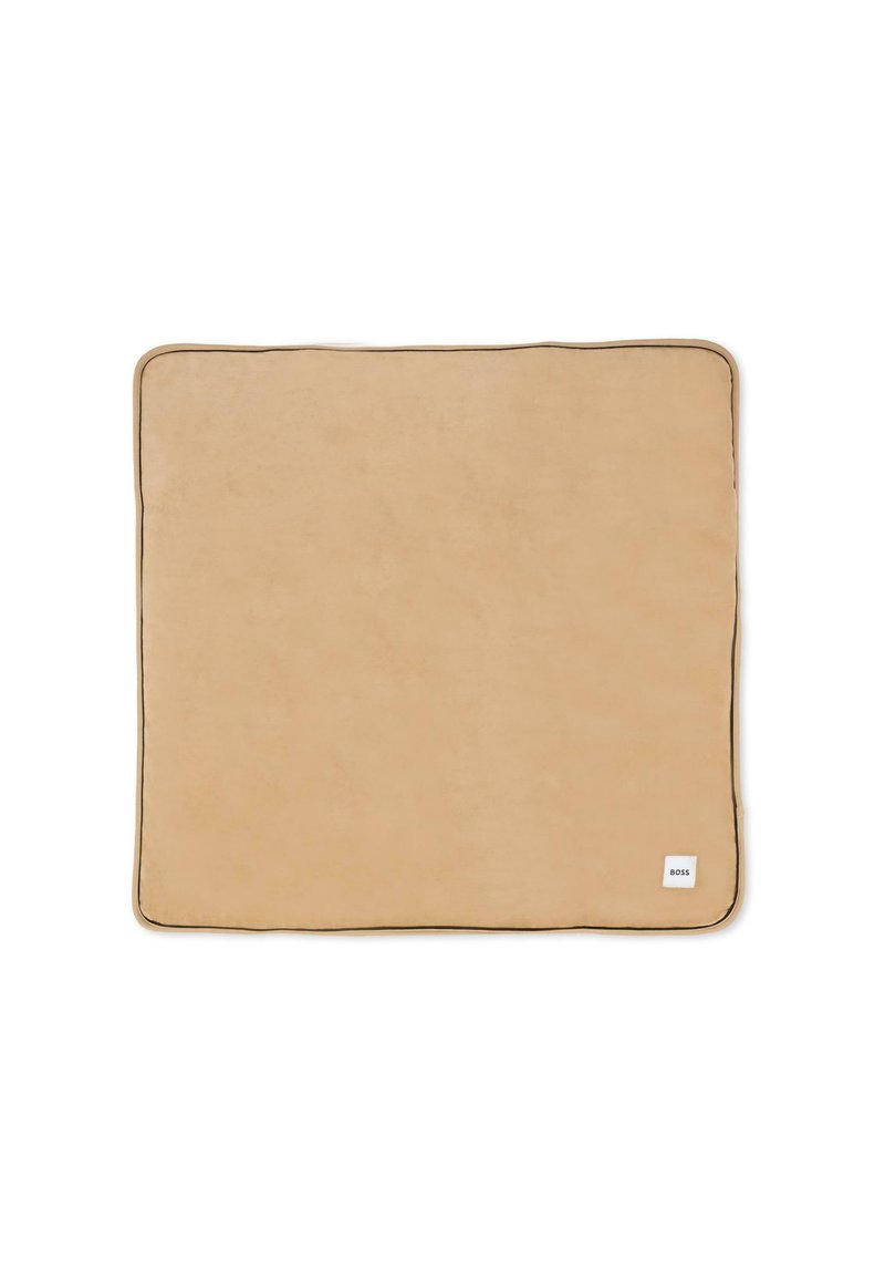 Beżowy prostokątny koc o miękkiej fakturze, wykończony kontrastowym ciemnym obszyciem. Na dole w rogu znajduje się mała biała etykieta.