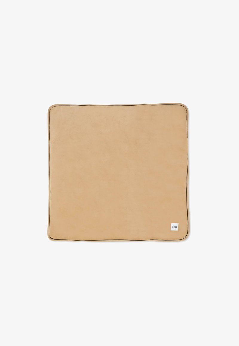 Beżowy prostokątny koc o miękkiej fakturze, wykończony kontrastowym ciemnym obszyciem. Na dole w rogu znajduje się mała biała etykieta.