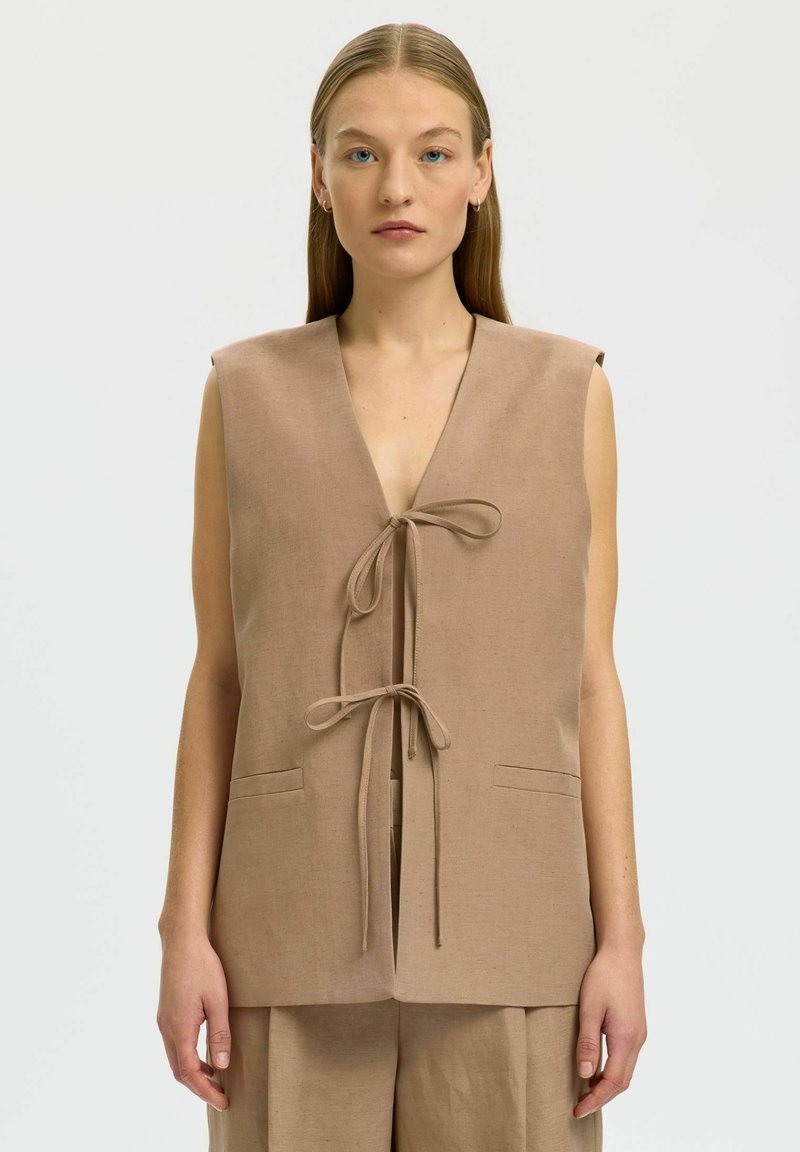 Femme aux longs cheveux blonds portant un gilet beige sans manches avec deux liens à l’avant et un pantalon beige assorti sur un fond uni.