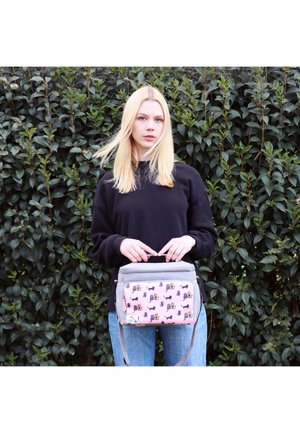 Junge Frau mit blonden Haaren in einem schwarzen Pullover, die eine grau-pinke Tasche mit Tiermotiven hält, steht vor einem dichten grünen Laubhintergrund.