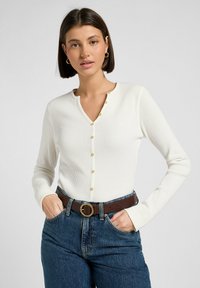 Top blanco de manga larga acanalada con escote redondo y botonera dorada, combinado con jeans azul de cintura alta y un cinturón marrón.